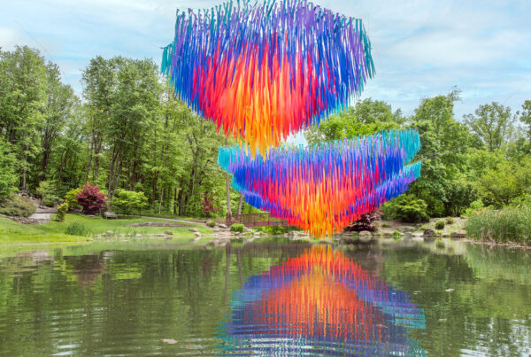 Two vibrant aerial sculptures from the Momentum art installation by Patrick Shearn of Poetic Kinetics suspended above a tranquil pond at Pittsburgh Botanic Garden, with colorful reflections mirrored in the water.